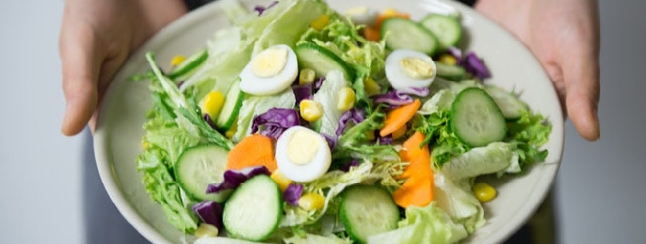 Hands holding out a keto friendly salad on a white plate. Salad has lettuce, cucumbers, hard-boiled eggs and no dressing.