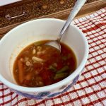 A close-up of a bowl of hearty vegetable soup with a silver spoon resting inside, placed on a red and white checkered cloth.